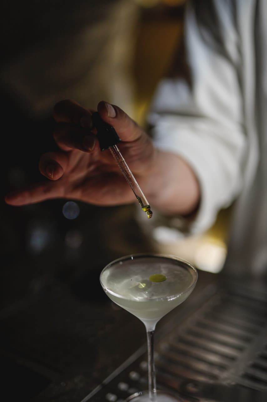 부산광역시 수영구 코듀로이 벨라즈 광안리 바 다찌석 혼술. 어두운 조명 아래 바텐더가 스포이드로 칵테일에 오일을 떨어뜨리는 순간. 투명한 액체와 오일 방울 질감이 강조된 1인석 현장.
