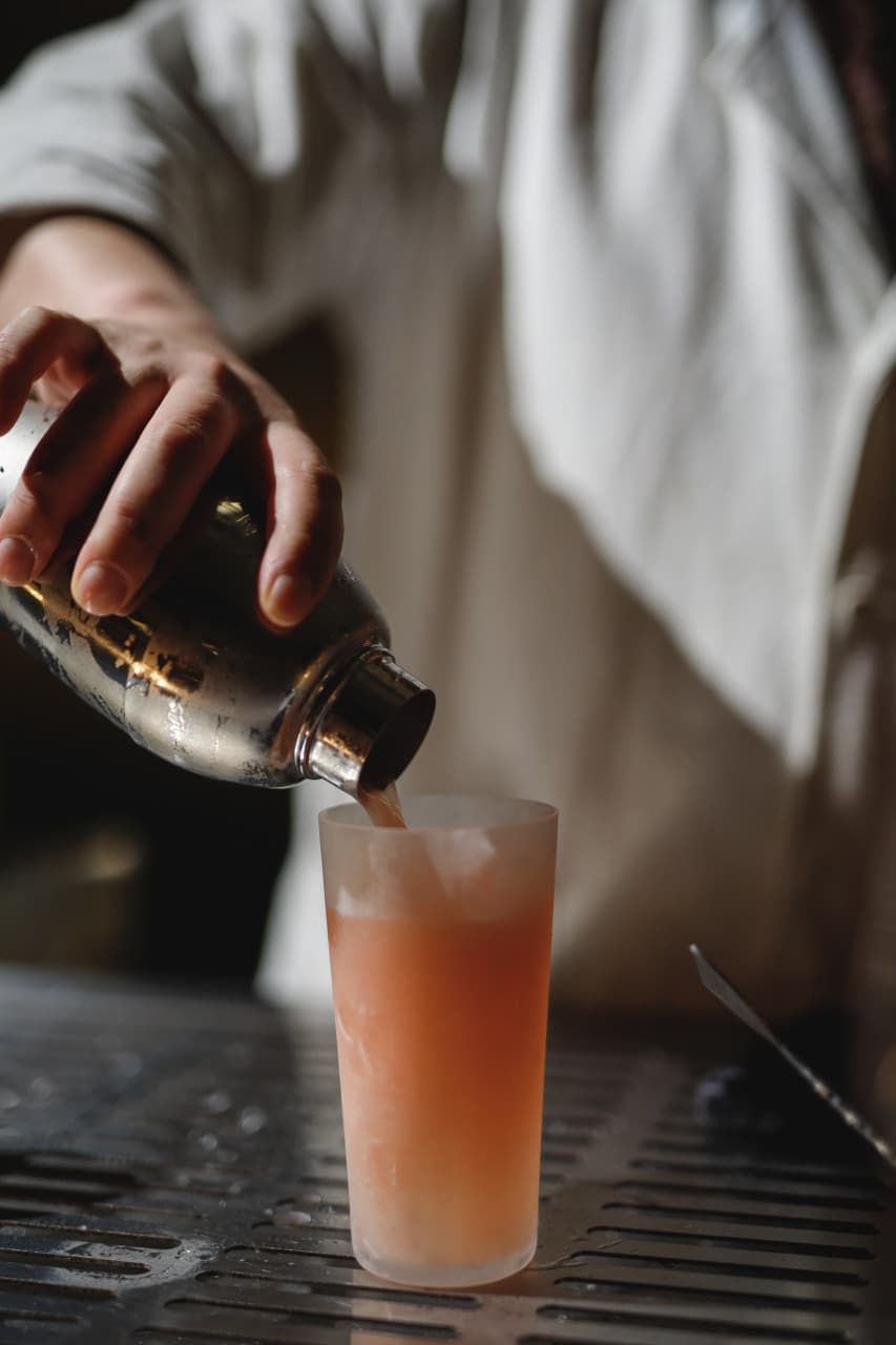 부산광역시 수영구 코듀로이 벨라즈 광안리 바(Bar) 다찌석에서 셰이커로 1인 칵테일을 따르는 상황. 은은한 조명 아래 핑크빛 액체와 얼음의 질감이 돋보이는 혼술 장면.