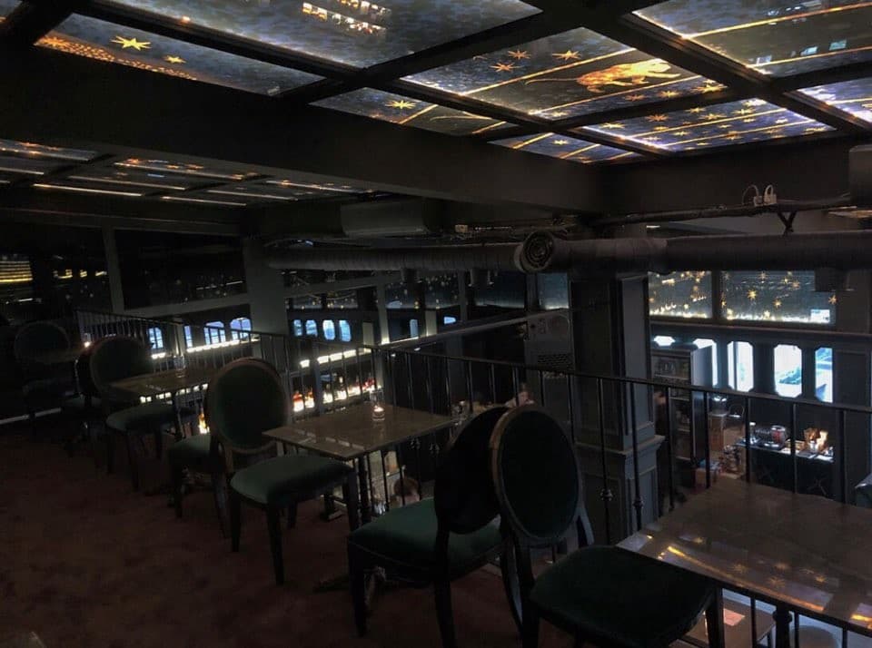 서울특별시 강남구 어글리 강남 바(Bar)의 별자리 패턴 천장 조명 아래 놓인 1인 테이블. 다찌석과 연결된 복층 난간 옆에서 혼술을 즐기기 용이한 좌석 배치를 보여줌.