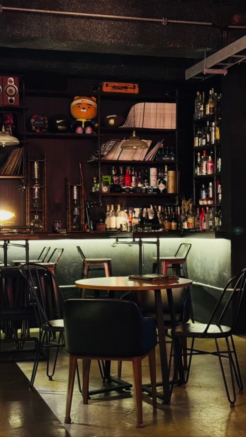 경기도 의정부시 유플레이온 바(Bar) 내부 다찌석과 원형 테이블. 선반 위 양주병과 LP판, 캐릭터 인형이 조명 아래 배치된 혼술 공간.