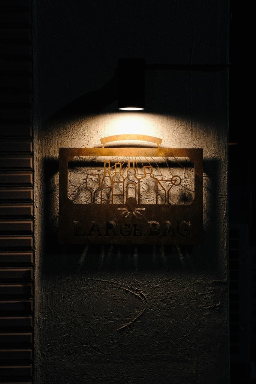 경상북도 경주시 라지백 경주 바(Bar) 입구. 상단 조명이 비추는 술병 실루엣 금속 간판과 거친 벽면. 1인 혼술 다찌석이 마련된 내부로 이어지는 외관.