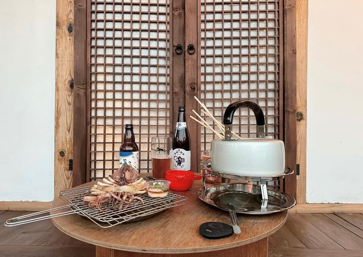 경상북도 경주시 황남주택 경주 맥주 호프. 한옥 격자문 앞 마루 위 원형 테이블에 차려진 구운 오징어와 주전자 오뎅탕. 1인 혼술에 적합한 야외 좌석과 병맥주가 놓인 구성.