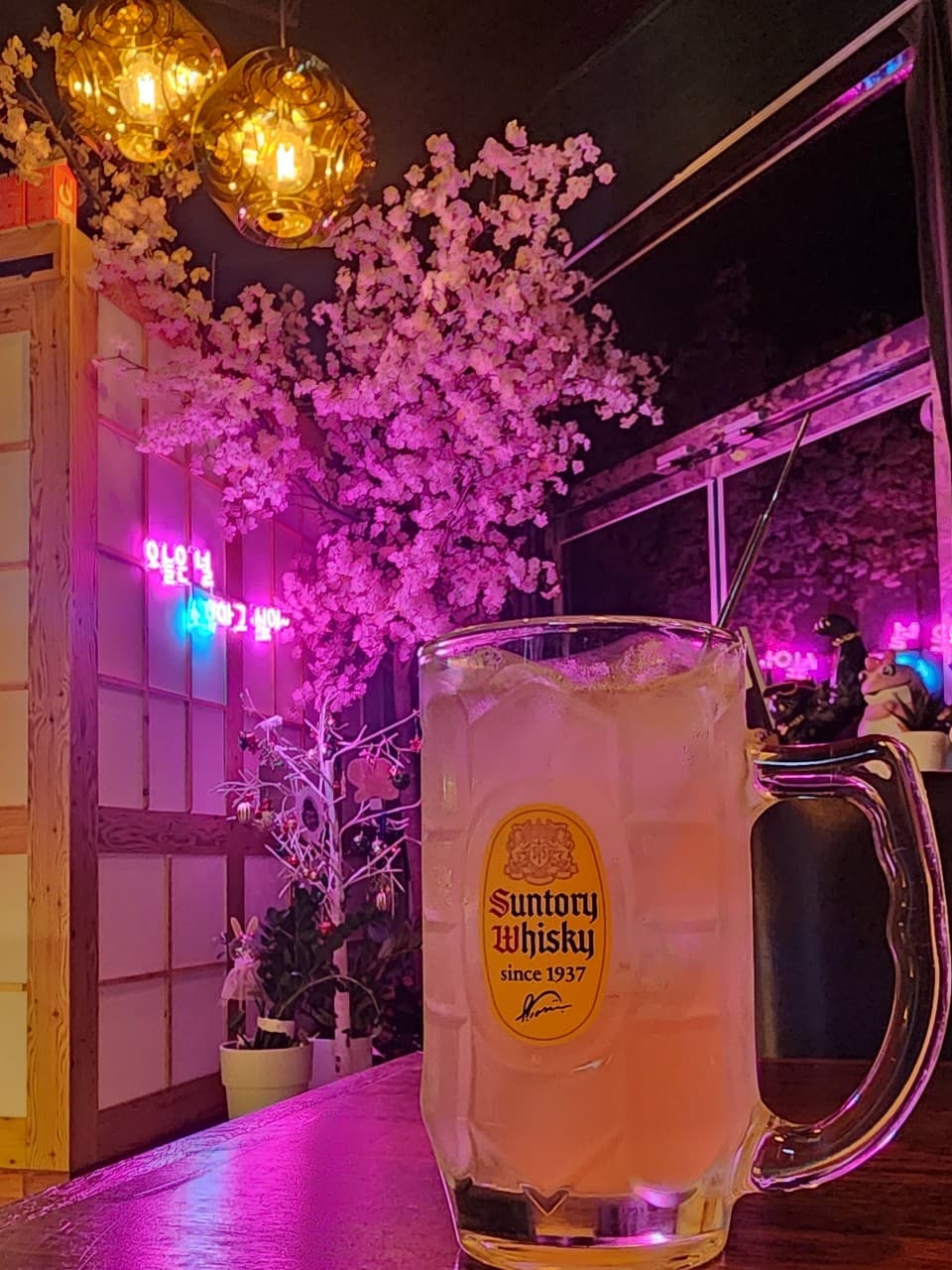 경상북도 포항시 소휴 포항 이자카야 다찌석 1인 혼술. 분홍색 네온 조명과 벚꽃 나무 아래 얼음이 담긴 산토리 하이볼이 놓인 목재 테이블 좌석.
