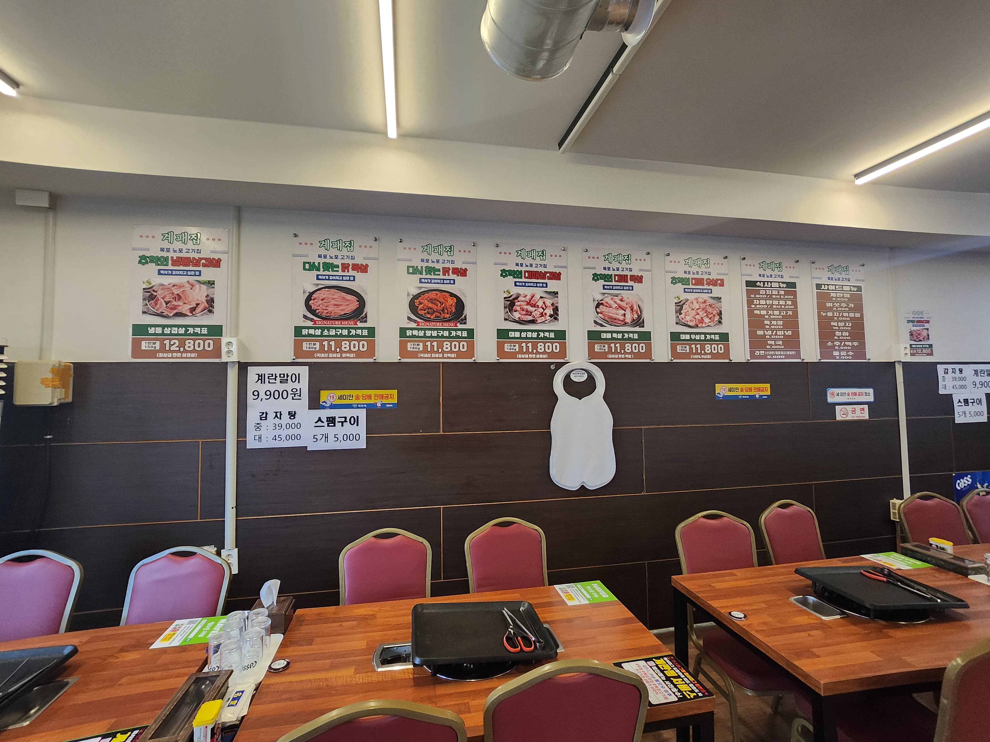 전라남도 목포시 목포 계패집 - 밝은 조명 아래 나무 테이블과 불판이 비치된 1인 혼밥 좌석. 선홍색 육질의 삼겹살 메뉴와 기타 식사 정보가 부착된 벽면과 앞치마가 보임.
