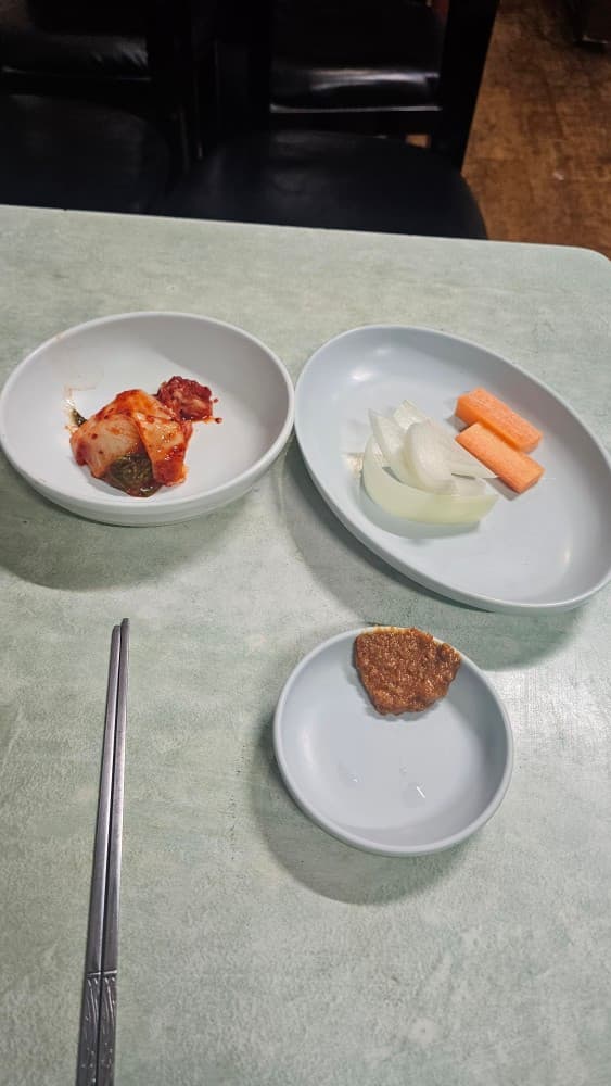 부산광역시 동구 솔잎국수 초량 포장마차 1인석 테이블 위 배추김치와 생양파, 당근, 쌈장이 차려진 모습. 밝은 조명 아래 쇠젓가락이 놓인 혼술 식사 상황