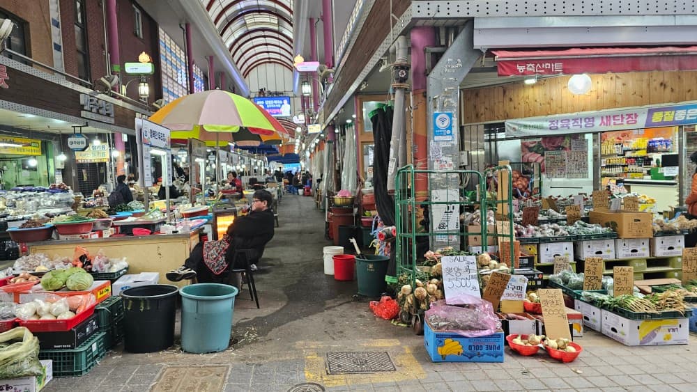 부산광역시 동구 신촌포차 초량 포장마차 - 전통시장 통로 파라솔 아래 1인석에 앉아 있는 모습과 주변에 진열된 채소 상자 및 식재료의 질감