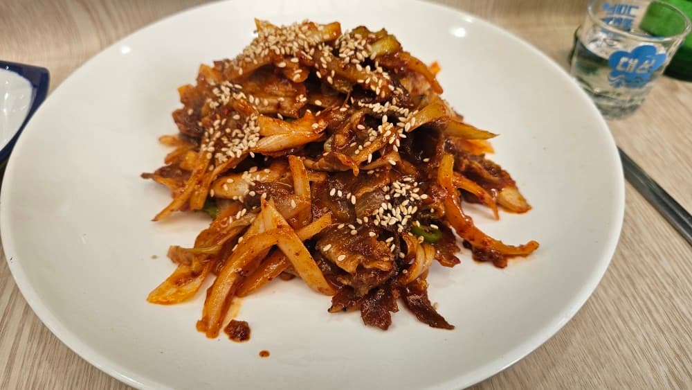 부산광역시 동구 신촌포차 초량 포장마차 다찌석에서 즐기는 1인 혼술. 깨를 뿌린 매콤한 제육볶음과 소주 한 잔이 차려진 나무 테이블의 정갈한 상황을 묘사함.