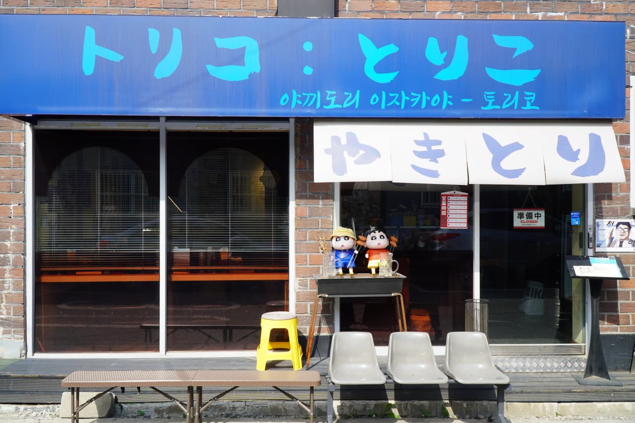 경기도 수원시 이자카야 토리코 파란 간판 아래 짱구 인형과 혼술 다찌석. 조명이 켜진 내부 1인석과 매장 앞 대기용 의자