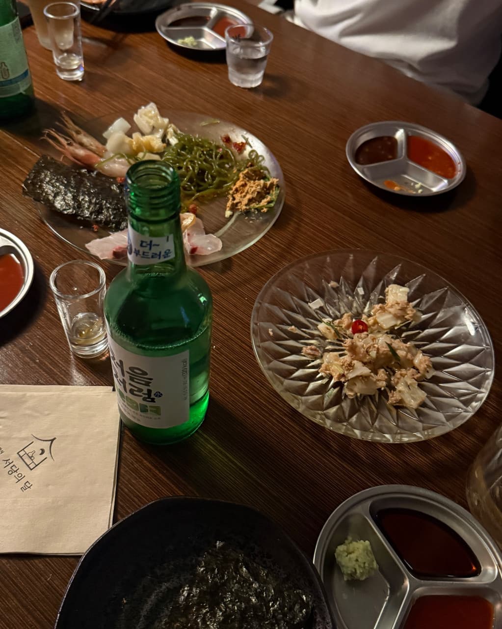경기도 부천시 요리주점 서당의달 나무 테이블 1인 다찌석 혼술. 투명한 접시에 담긴 사시미와 해초, 소주와 소스 그릇이 차려진 상차림 위로 떨어지는 주황빛 조명.