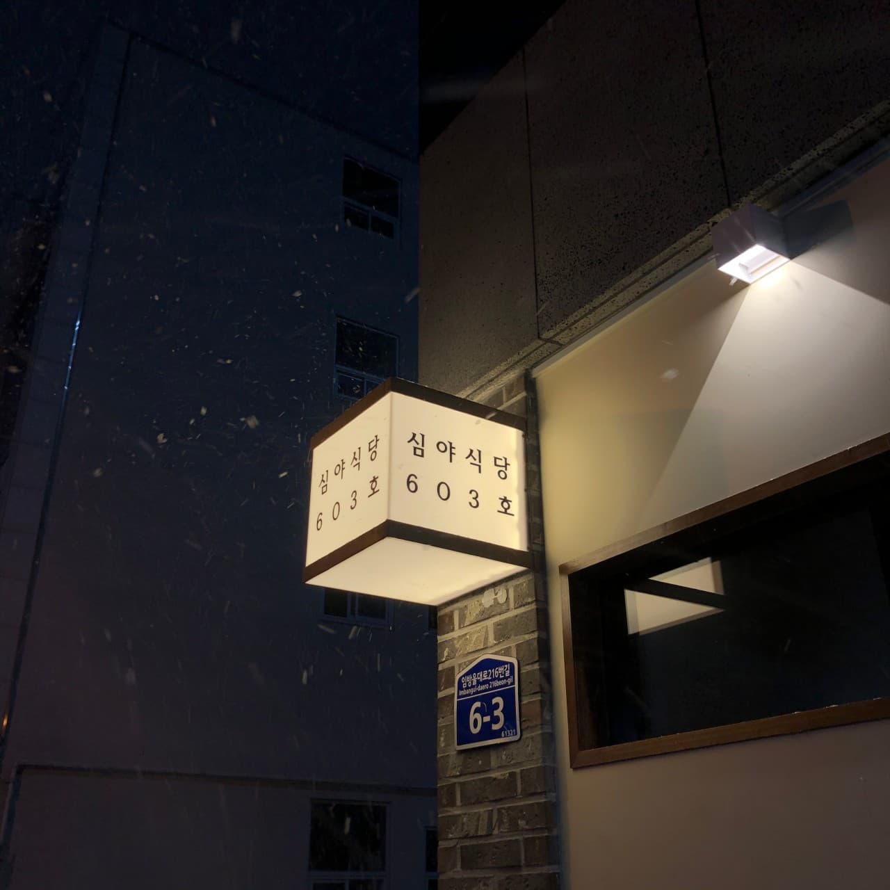 광주광역시 광산구 심야식당 603호 광주 이자카야. 눈 내리는 밤 조명 켜진 정육면체 간판과 혼술 손님을 맞이하는 건물 외벽 전경.