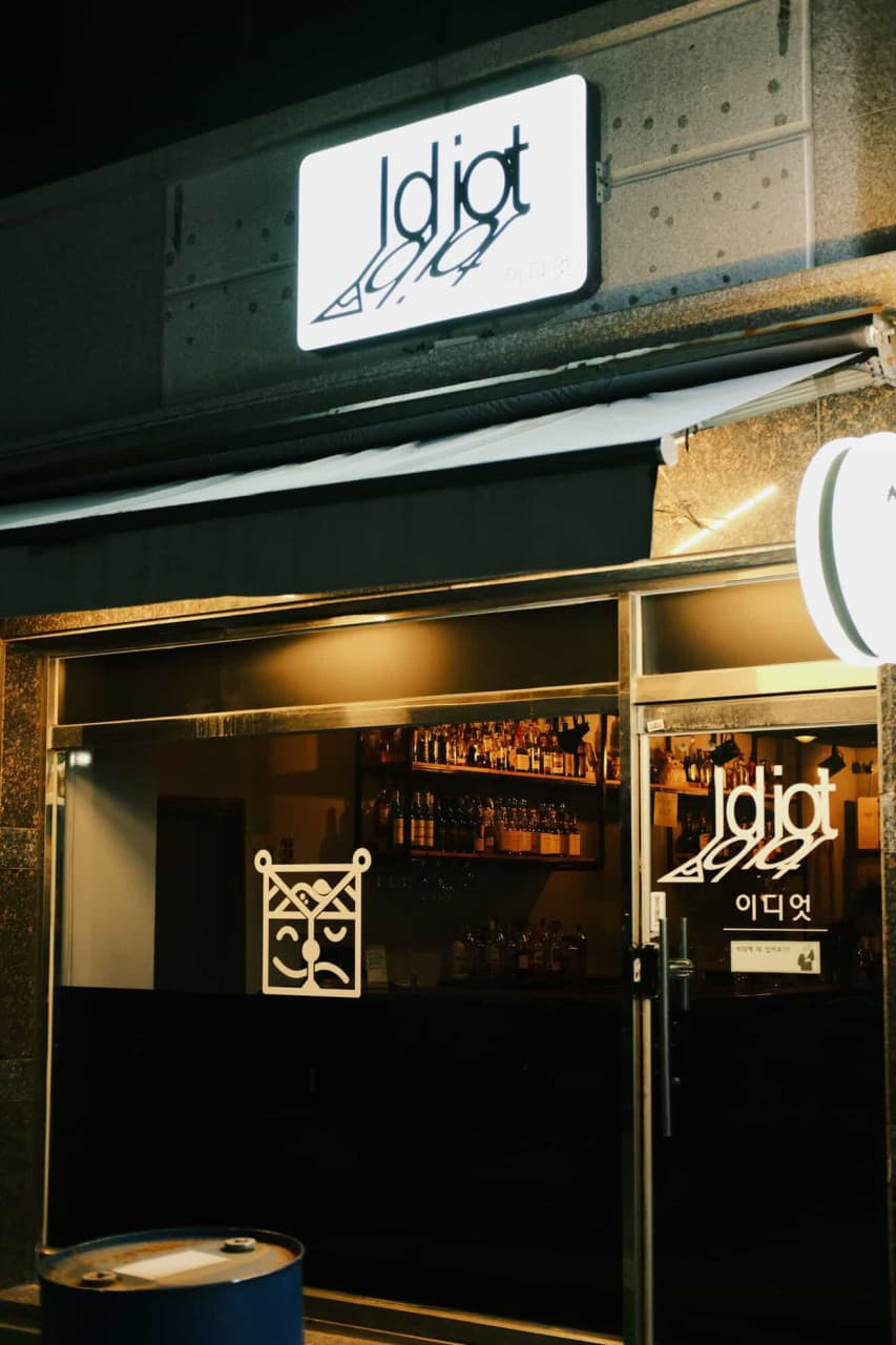 광주광역시 광산구 이자카야 이디엇 광주 입구. 화이트 사인 보드 아래 노란 조명이 비치는 실내에는 다양한 술병과 혼술 전용 다찌석이 보임.