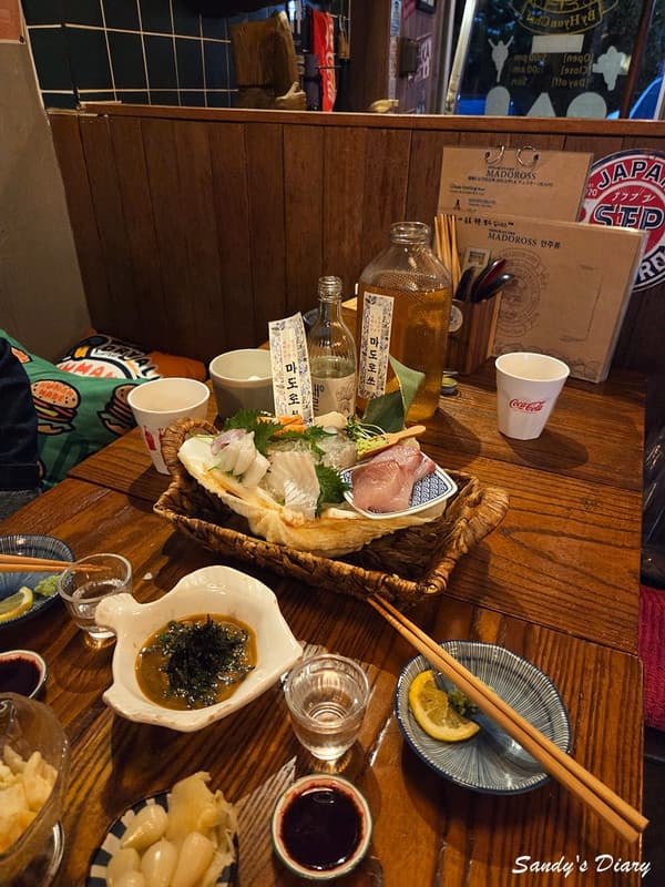 경기도 광주시 이자카야 마도로쓰 목재 다찌석에 차려진 1인 사시미와 혼술 상차림. 낮은 조도의 조명 아래 신선한 회의 질감과 전용 술병이 놓인 카운터 좌석의 모습.