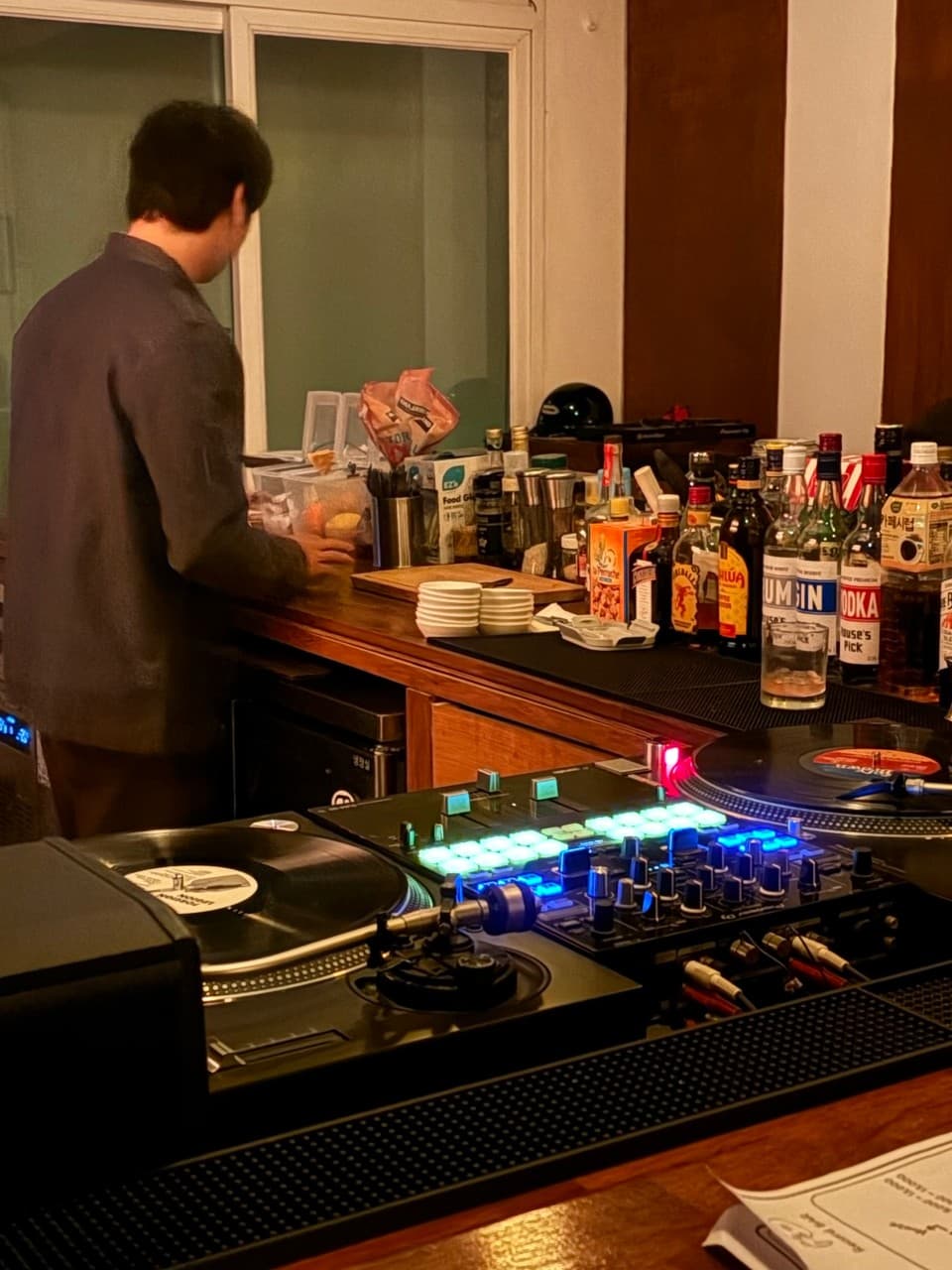 경기도 광주시 섭레코드바 LP바의 주황빛 조명 아래 다찌석. LP 턴테이블과 믹서, 진과 보드카 병이 나열된 나무 카운터에서 바텐더가 혼술 손님을 위한 안주를 준비하는 상황.