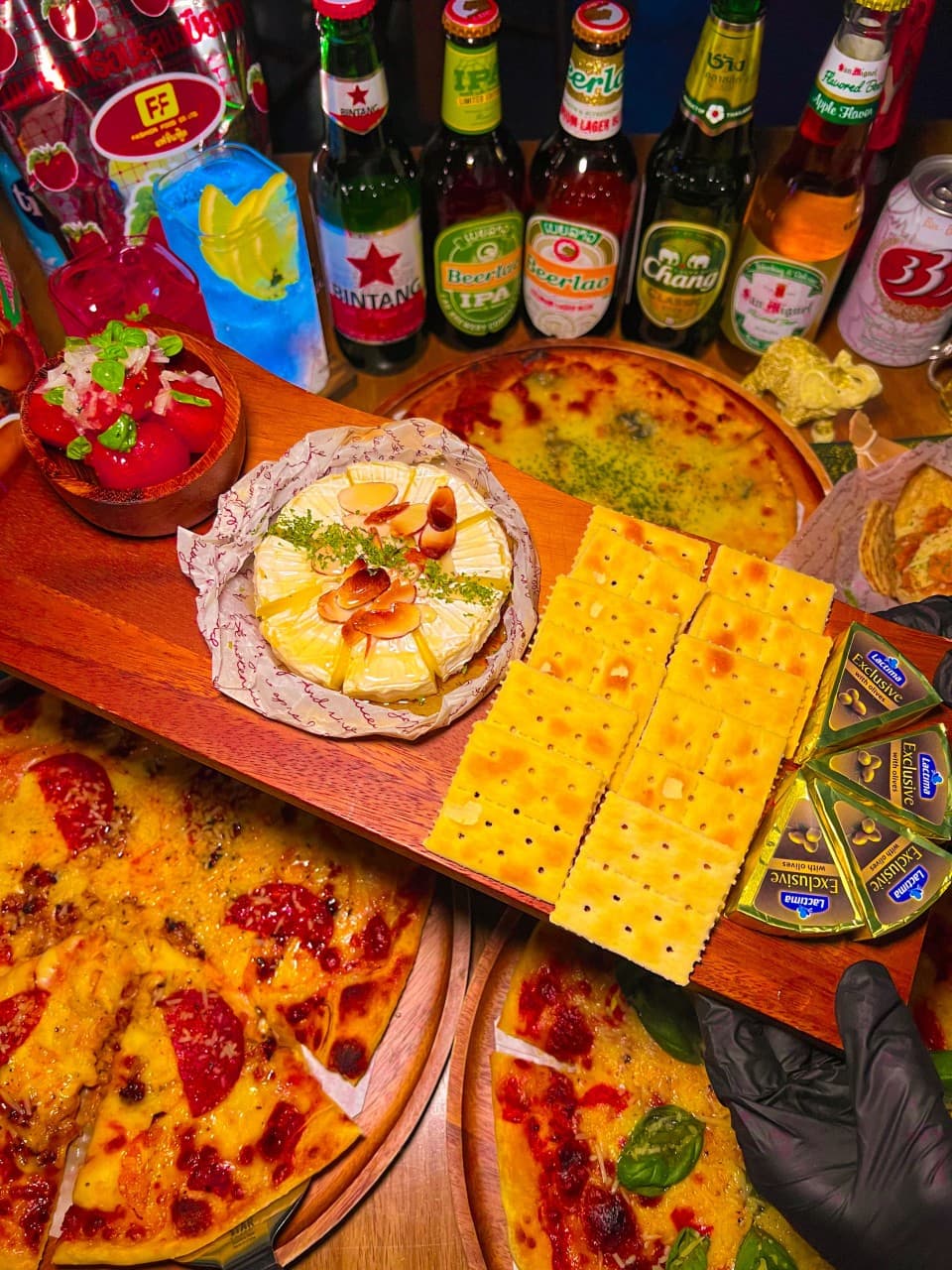 대구광역시 중구 맥주 호프 싸눅사운드 대구 - 노란 조명 아래 페퍼로니 피자와 견과류를 올린 브리치즈, 세계 맥주가 차려진 1인 다찌석. 혼술을 위한 안주 세트와 색감이 돋보이는 칵테일 구성