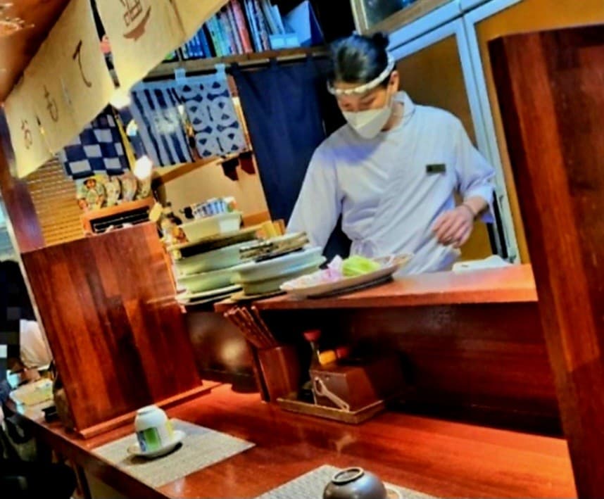 대구광역시 수성구 이자카야 오모테 대구의 1인 다찌석. 일본식 노렌이 걸린 나무 카운터에서 셰프가 음식을 준비하며 조명이 식기를 비추는 혼술 상황을 보여줌.
