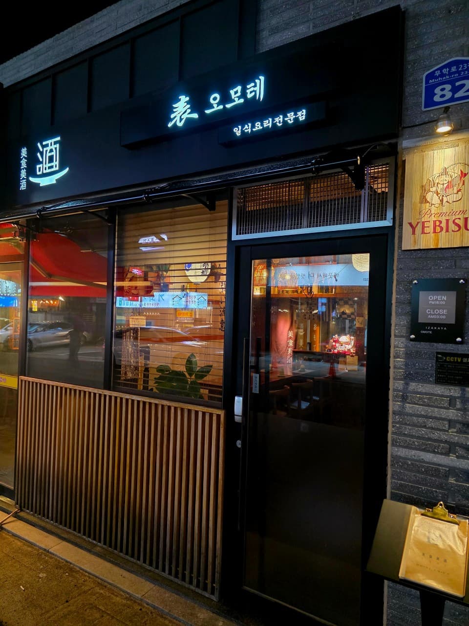 대구광역시 수성구 이자카야 오모테 대구 - 조명이 켜진 간판과 나무 간살, 유리창 너머로 1인 다찌석이 보이는 혼술 공간의 입구.
