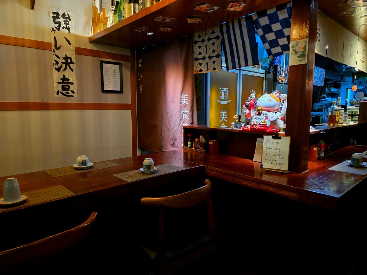 대구광역시 수성구 이자카야 오모테 대구 목재 다찌석에 차려진 1인 식기. 전구색 조명과 마네키네코 인형, 서예 족자가 배치된 내부에서 혼술을 즐기는 상황.
