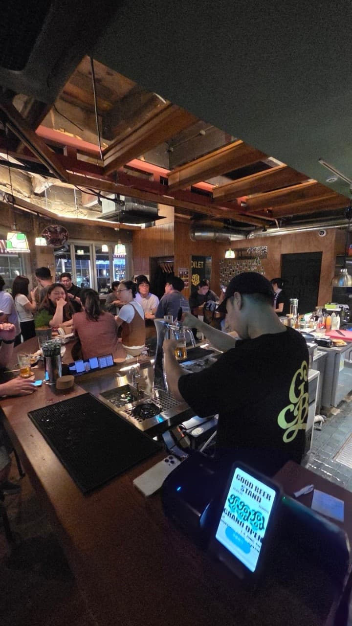 대구광역시 중구 굿비어스탠드 대구 맥주 호프 매장. 황색 조명 아래 곡선형 나무 다찌석과 1인석에서 혼술을 즐기는 사람들과 직원이 맥주를 따르는 상황.