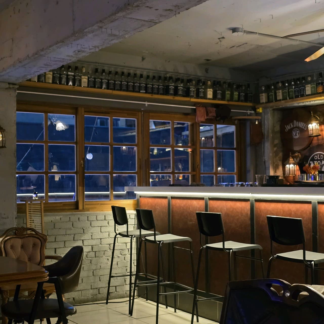 대구광역시 중구 헬로외국포차 대구 맥주 호프 - 하단 조명이 비치는 다찌석과 격자창 밖 야경이 보이는 혼술 공간. 상단 선반을 채운 위스키 병들과 거친 질감의 노출 콘크리트 인테리어.