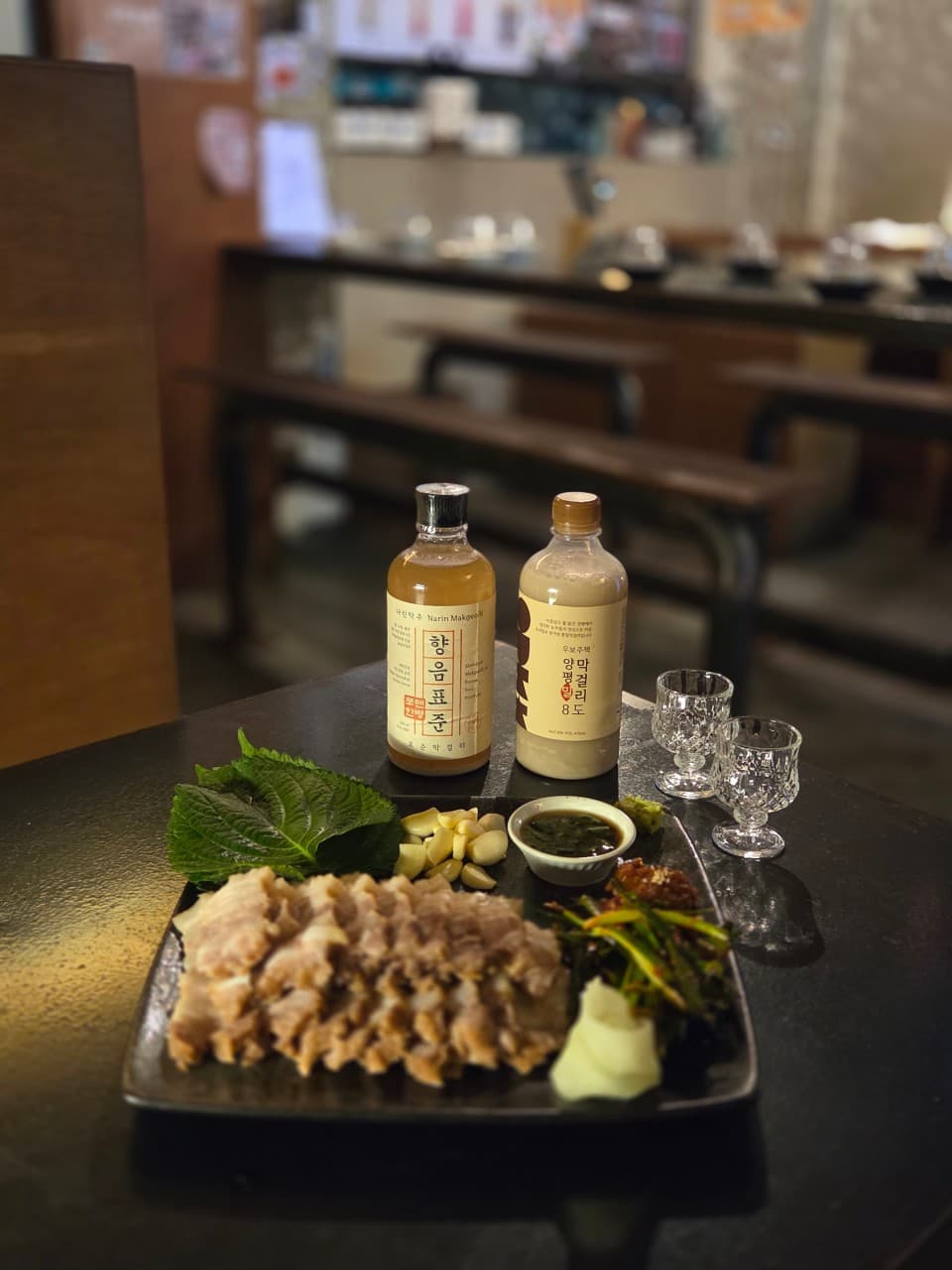 대구광역시 중구 전통주점 활력의 노란 조명 아래 차려진 1인 보쌈과 전통주. 어두운 다찌석 테이블 위 수육의 질감과 막걸리 병이 나란히 배치된 혼술 상황.