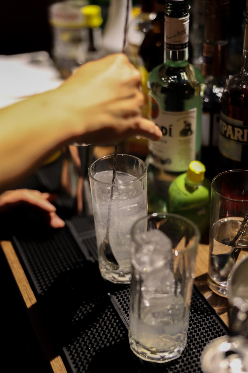대구광역시 중구 혼술바 결 - 바(Bar) 다찌석에서 바텐더가 1인용 하이볼을 젓는 모습. 핀 조명 아래 얼음이 담긴 유리잔과 다양한 양주병이 배치됨.