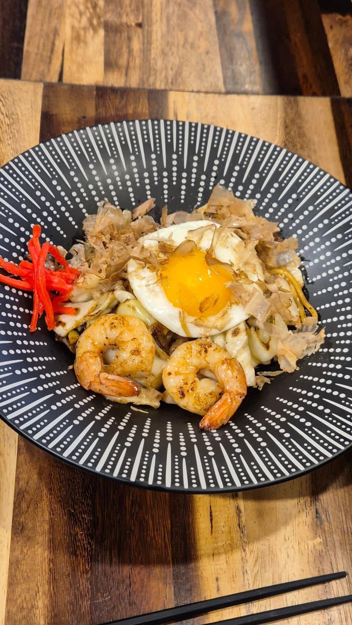 전남광주통합특별시 순천시 야키토리 신지야 순천 이자카야 다찌석. 나무 테이블 위 조명을 받아 윤기 나는 새우와 계란 노른자가 올라간 1인 볶음면이 차려진 혼술 분위기.
