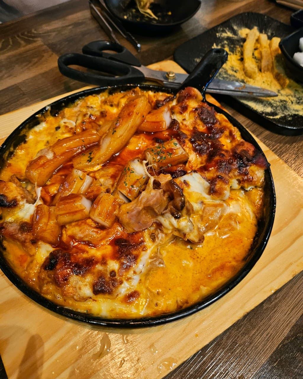 인천광역시 검단구 가마꿉 당하 본점 인천의 1인 다찌석 혼술. 무쇠 팬에 가득 담긴 치즈 치킨,닭강정과 떡사리. 따뜻한 조명 아래 목재 테이블 위에서 집게와 가위로 식사하는 상황.