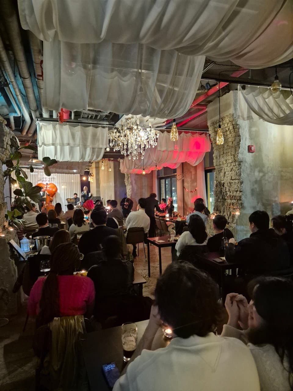 인천광역시 연수구 재즈257 송도센트럴파크점 인천 와인 바의 샹들리에 아래, 1인 좌석에서 공연을 감상하는 혼술 상황. 테이블 위 와인 잔과 흰 천 장식이 어우러진 무대 앞 전경.
