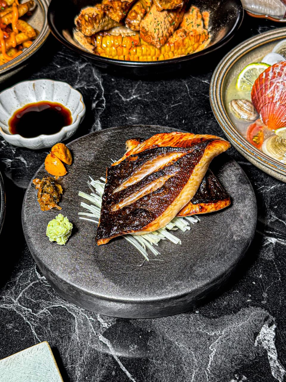 인천광역시 남동구 이자카야 물고기골목 인천의 다찌석 혼술 차림. 검은 돌판 위 노릇하게 구워진 생선구이와 와사비, 가리비 술찜과 옥수수 튀김의 질감이 조명 아래 선명함.