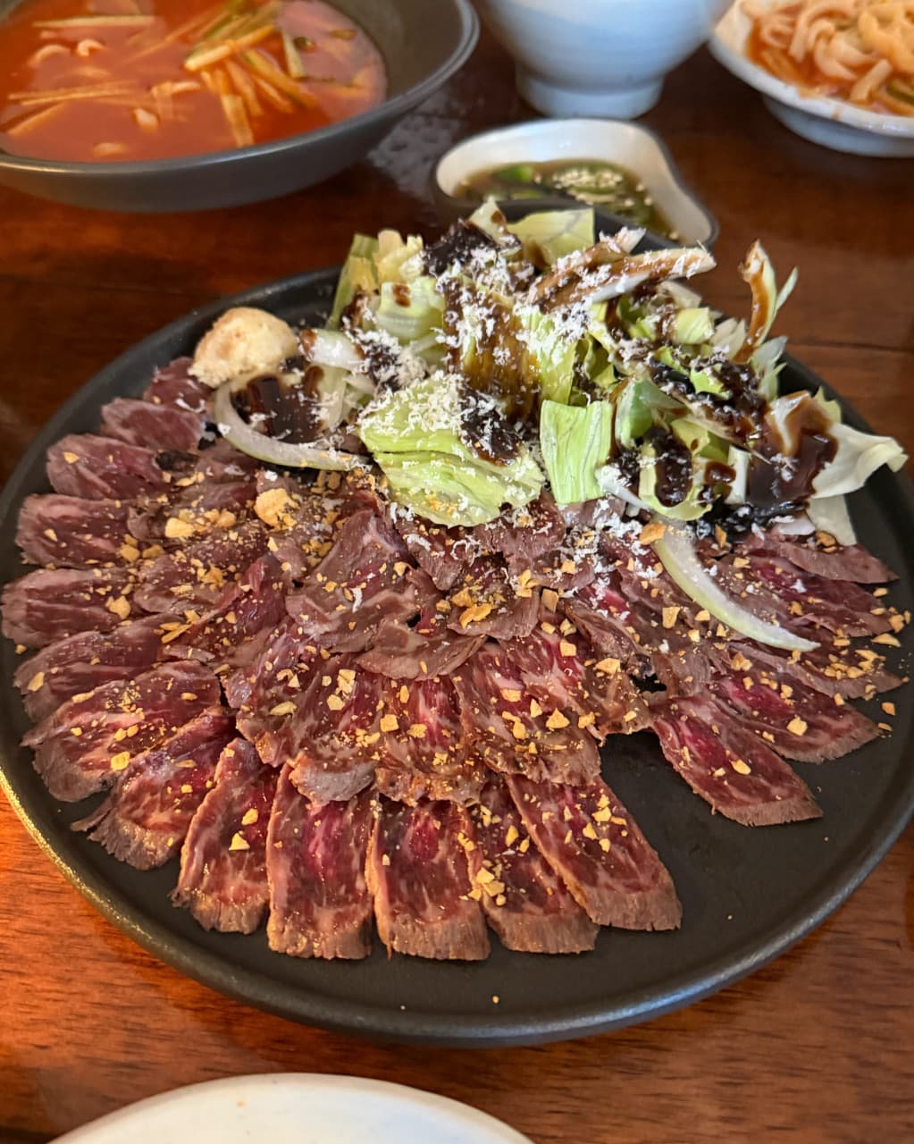 인천광역시 계양구 요리주점 저녁산책 인천 다찌석 혼술 메뉴 소고기 타타키. 나무 테이블 위 조명 아래 놓인 선홍빛 육질과 마늘 칩, 채소 샐러드 구성.