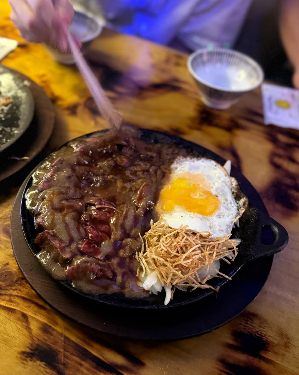 인천광역시 남동구 혼술 이자카야 코코스키 간석점 인천 - 나무 다찌석 위 조명을 받은 철판 스테이크와 계란후라이. 소스의 윤기와 튀김 고명의 질감이 선명하게 보이는 1인 메뉴.