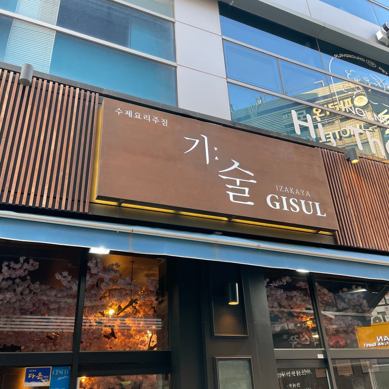 경기도 안산시 요리주점 기술 경기 안산의 갈색 간판과 나무 살 외벽. 창 안쪽 분홍색 벚꽃 조화와 노란 조명이 비치는 혼술 하기 적합한 매장 외부 전경.