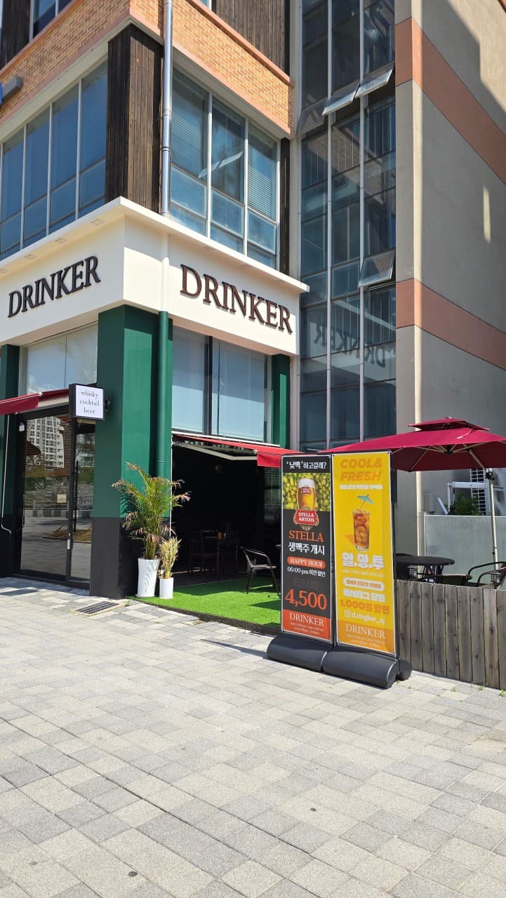 세종특별자치시 보듬3로 드링커 세종 바(Bar). 흰색 간판 아래 초록색 벽면과 테라스 인조잔디 위로 1인 혼술용 야외 좌석과 레드 파라솔, 주류 홍보 배너가 설치됨.