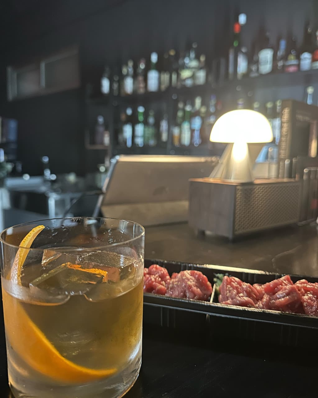 세종특별자치시 조치원읍 비버 세종 바(Bar) 다찌석 혼술. 오렌지 껍질을 넣은 칵테일과 육회가 차려진 1인 테이블 위로 버섯 조명과 선반 가득한 술병들이 위치함.