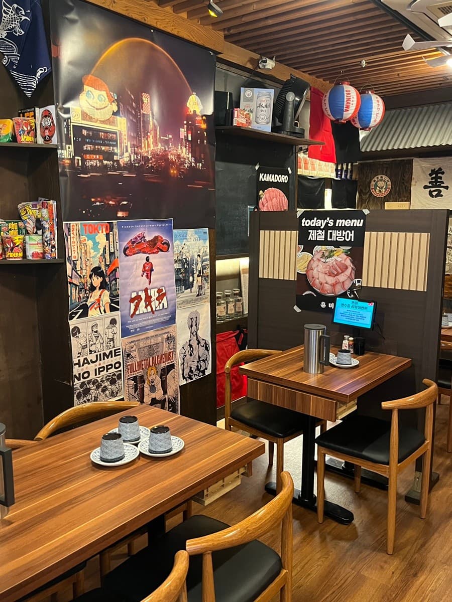 세종특별자치시 보듬3로 이자카야혼마 세종 이자카야. 일본 애니메이션 포스터가 붙은 벽면 아래 제철 대방어 메뉴가 안내된 1인 혼술용 나무 테이블과 좌석이 위치한 내부 공간.