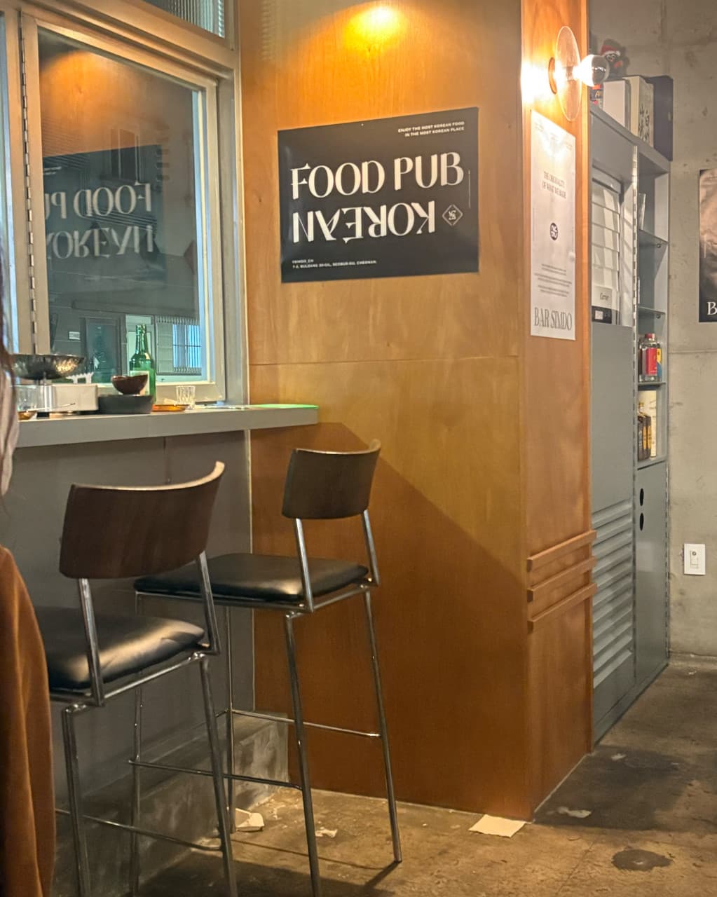 대전광역시 유성구 이자카야 심도 봉명점 대전의 주황색 조명이 비추는 다찌석과 높은 의자. 벽면의 포스터와 창가 테이블에 놓인 술병이 혼술 분위기를 조성함.