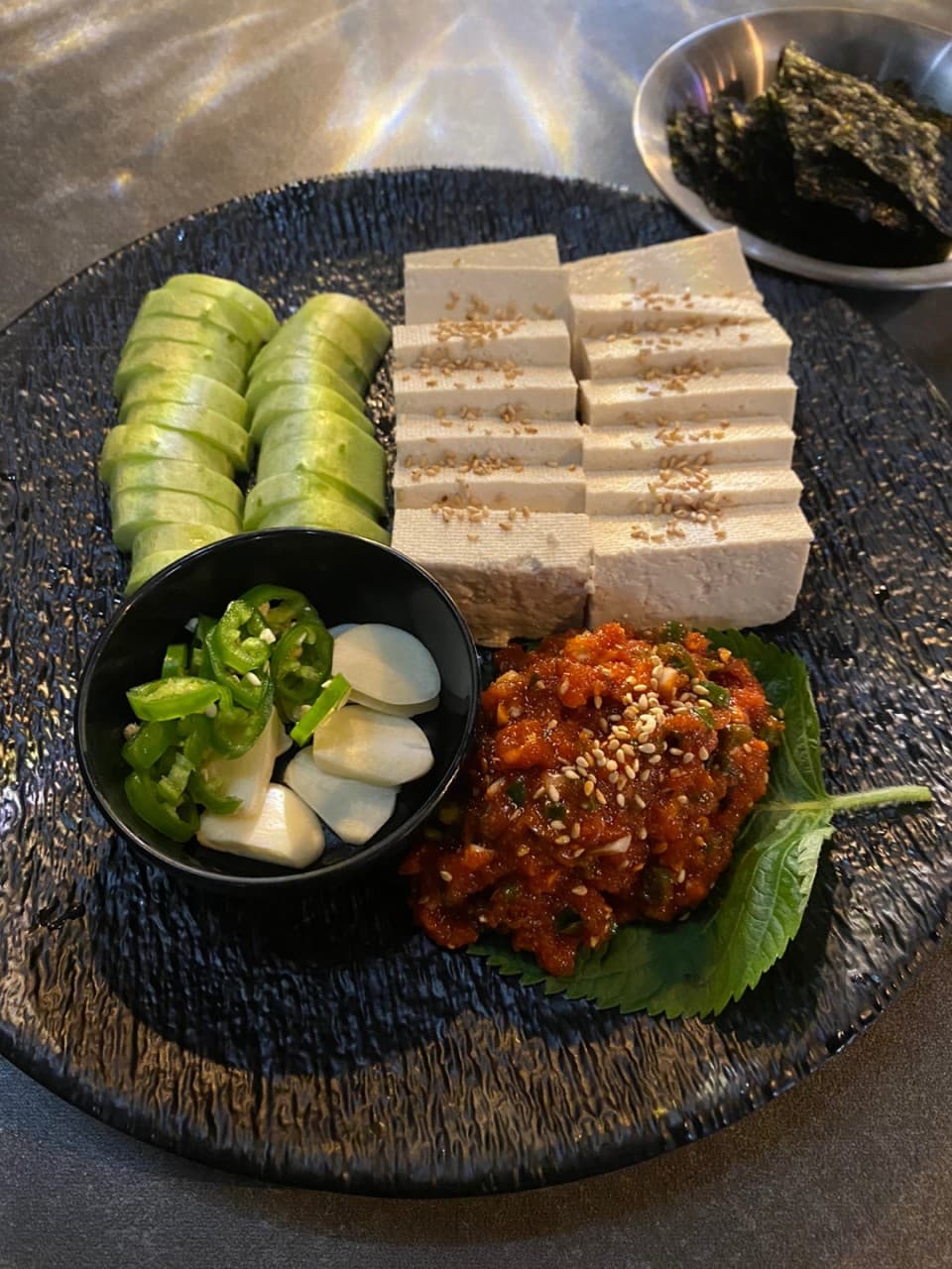 대전광역시 서구 화목식탁 대전 요리주점 다찌석 조명 아래 놓인 1인 혼술 안주. 검은 접시 위 나란히 놓인 두부와 오이, 양념 명란, 마늘과 고추, 김이 어우러진 차림.