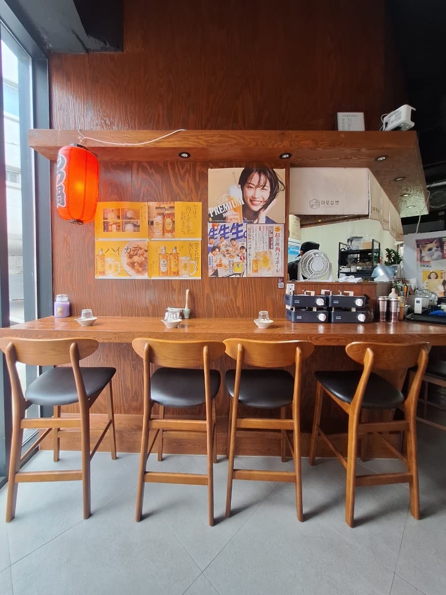 대전광역시 서구 마포십번 갈마본점 이자카야 붉은 제등 아래 우드톤 다찌석. 벽면의 하이볼 포스터와 1인 의자가 배치된 혼술 상황의 매장 내부 구성.