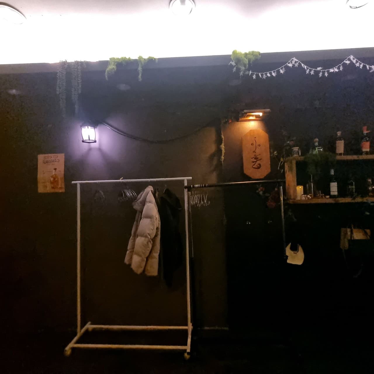 인천광역시 남동구 제주아홉 혼술바 인천구월점 인천 바(Bar) - 어두운 벽면 조명과 외투 행거, 술병이 진열된 선반이 있는 1인 혼술객을 위한 내부 공간