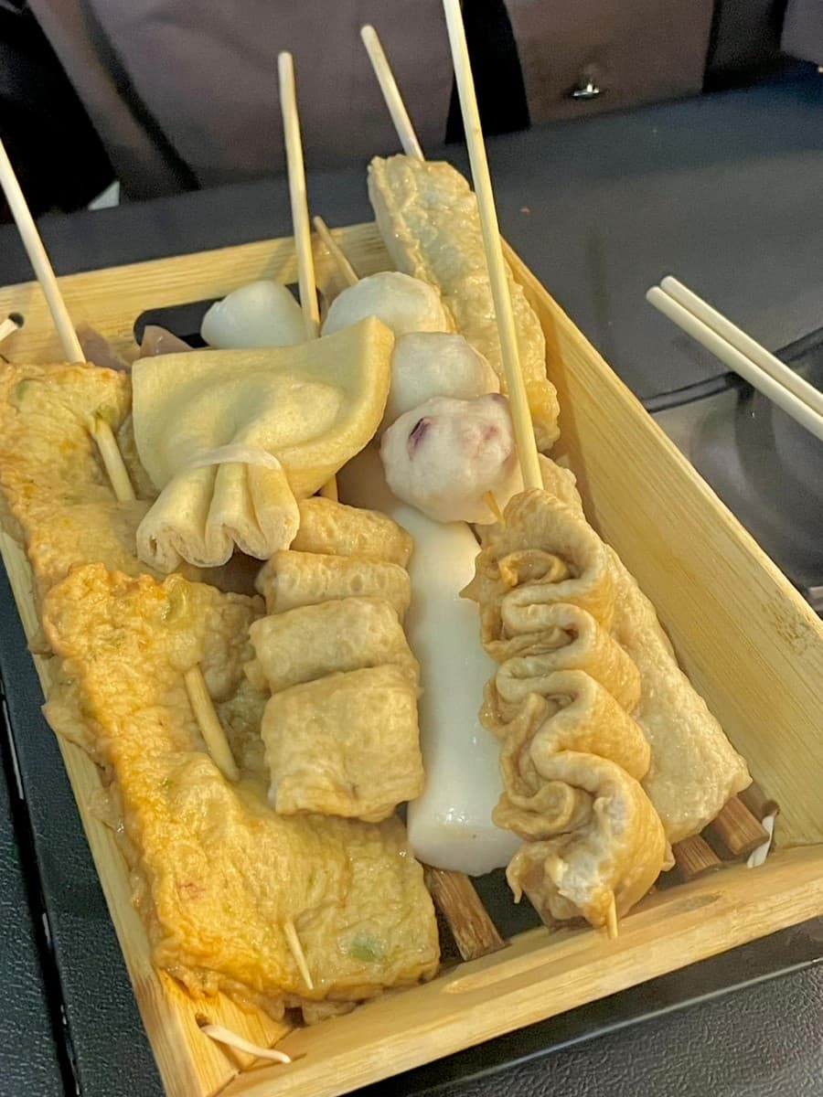 인천광역시 남동구 요리주점 철길부산집 인천구월점 인천 - 조명 아래 1인 다찌석 혼술 어묵 모듬과 나무 용기에 담긴 유부주머니, 물떡 꼬치의 질감이 보이는 분위기