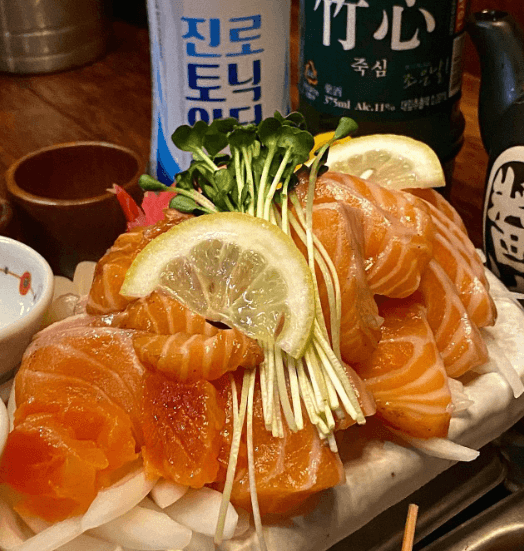 인천광역시 남동구 요리주점 철길부산집 인천구월점 인천 - 조명 아래 다찌석 1인 연어 사시미와 주류가 놓인 혼술 상황