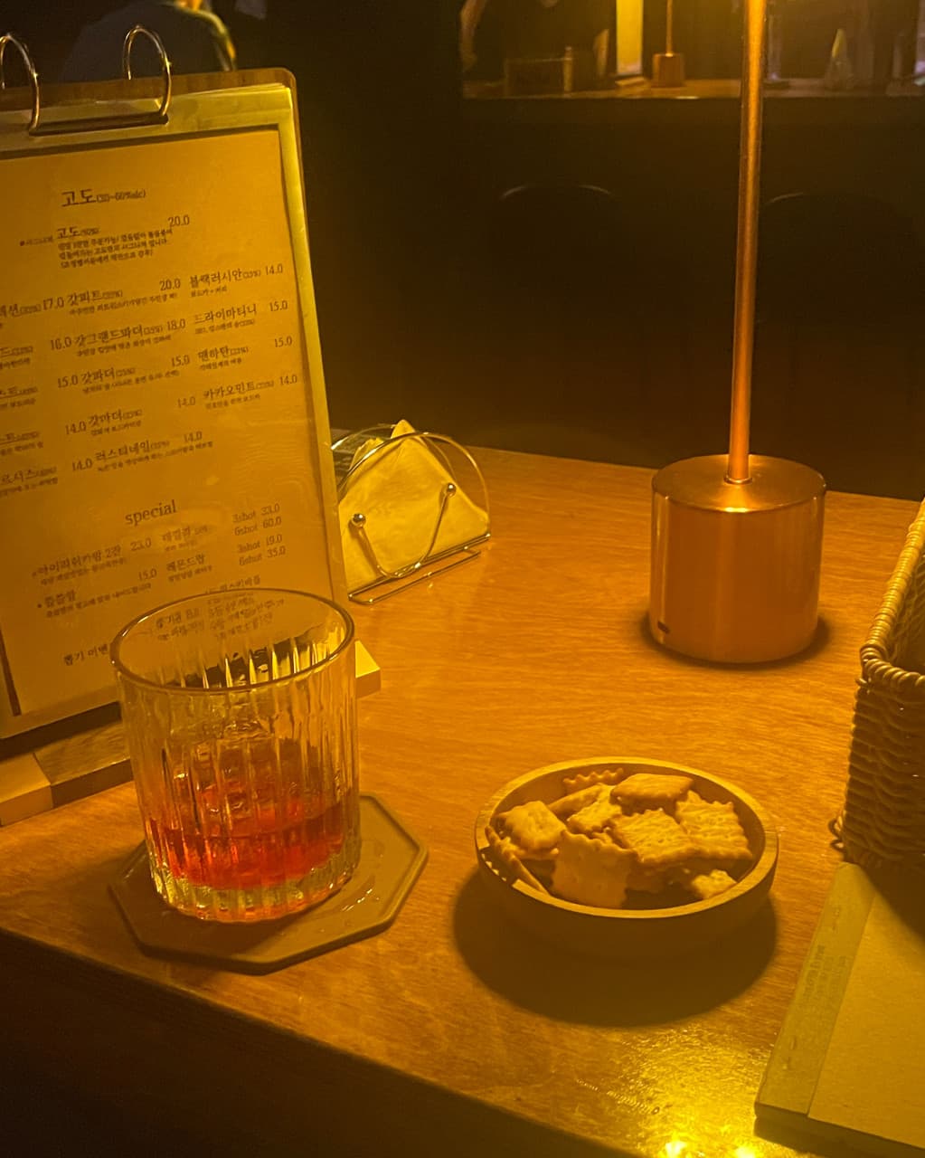 인천광역시 부평구 혼술바 고도 부평점 인천의 노란 조명 아래 1인 다찌석. 위스키 잔과 바삭한 크래커가 놓인 목재 테이블 위 메뉴판과 금색 스탠드가 배치된 바(Bar) 혼술 상황.