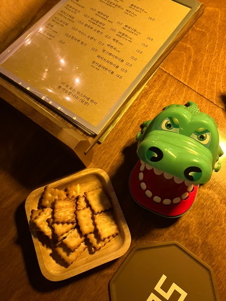 인천광역시 부평구 혼술바 고도 부평점 인천 바(Bar) 다찌석의 노란 조명 아래 놓인 메뉴판과 나무 접시 위 바삭한 크래커, 악어 장난감으로 구성된 혼술 공간