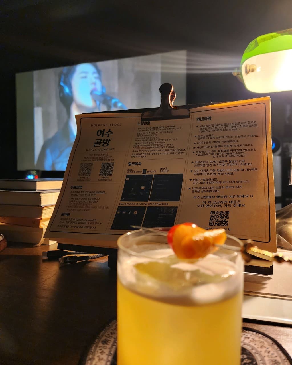 전남광주통합특별시 여수시 여수골방 전남 바(Bar) 1인 다찌석. 초록색 스탠드와 대형 스크린 배경 속 과일 가니쉬를 올린 노란색 칵테일 한 잔이 놓인 혼술 상황.