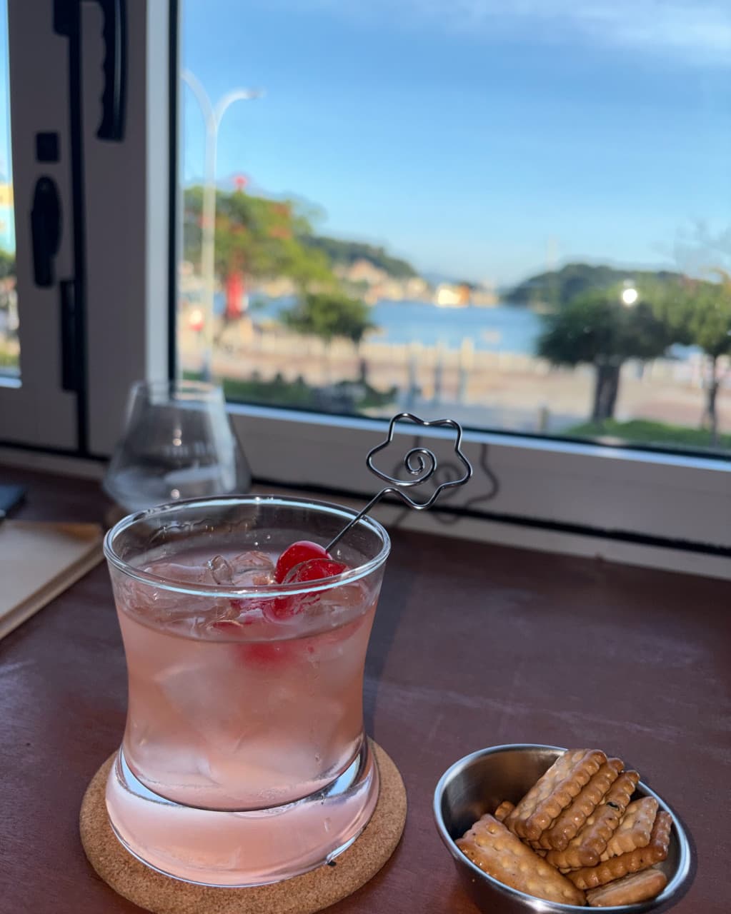 전남광주통합특별시 여수시 등대이층 전남 바(Bar) 창가 1인 다찌석. 자연광 아래 분홍색 칵테일과 체리, 크래커가 차려진 모습. 바다와 등대 풍경 배경의 혼술 상황.