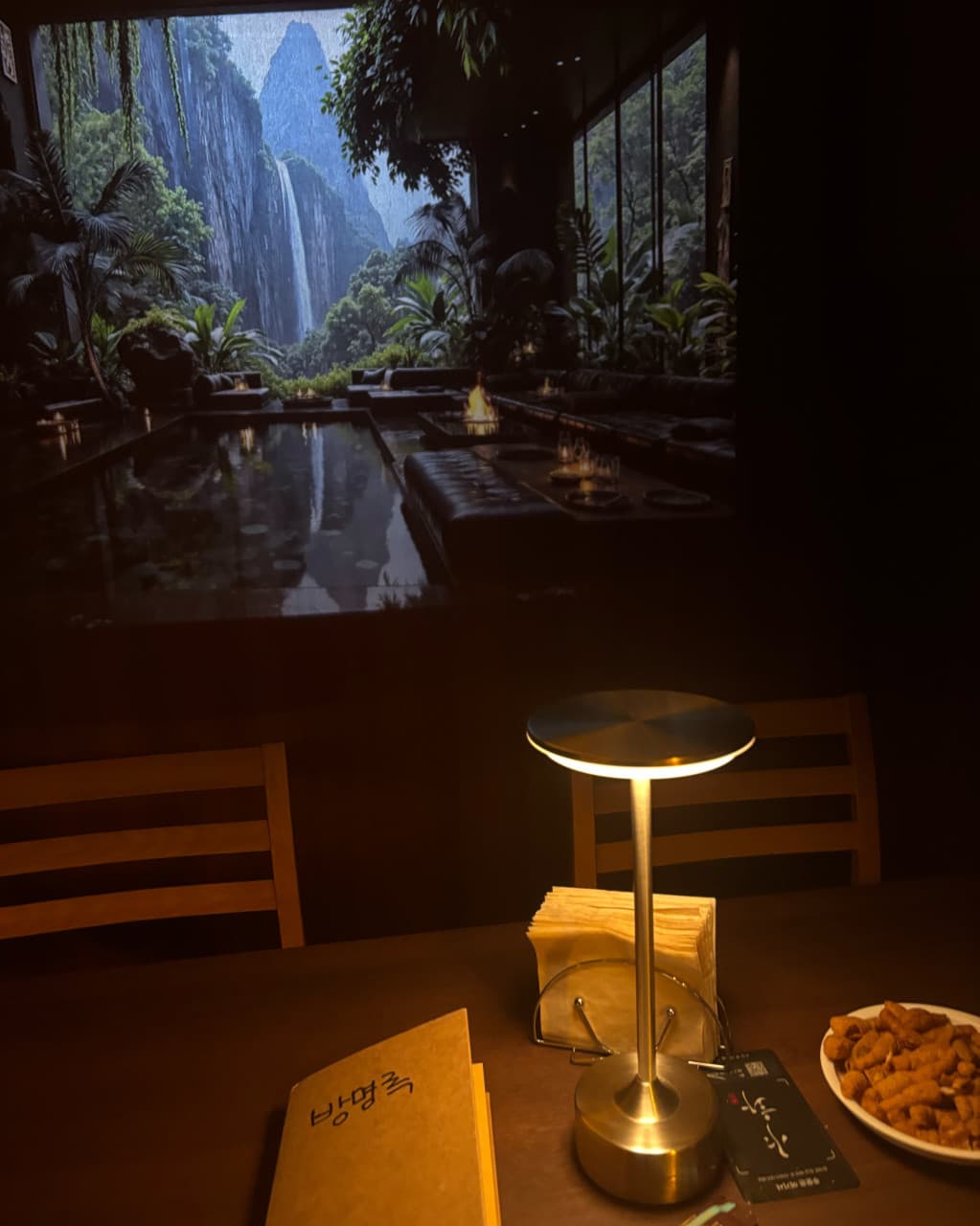 전남광주통합특별시 서구 야화 혼술바 광주상무점 전남 바(Bar) 1인석의 스탠드 조명 아래 놓인 기본 안주와 방명록. 배경의 대형 폭포 미디어아트가 보이는 혼술 자리.
