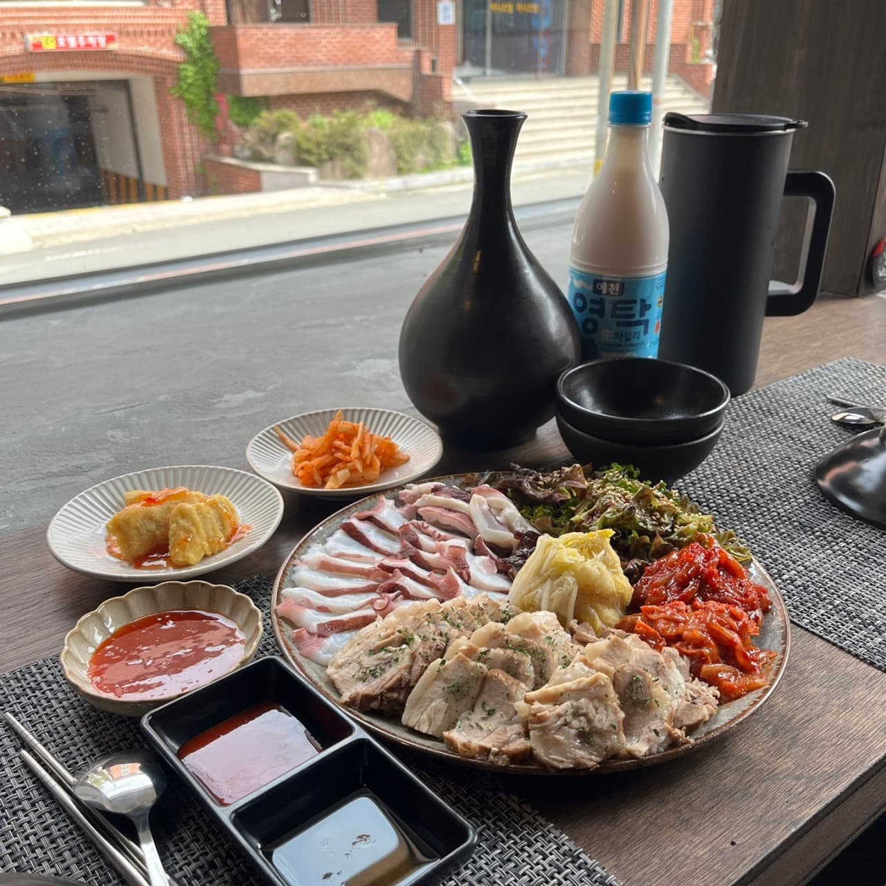 경상북도 경주시 안다미로 경북 요리주점 창가 다찌석 혼술. 자연광 아래 보쌈과 문어숙회, 막걸리가 차려진 회색 매트 위 1인 상차림.