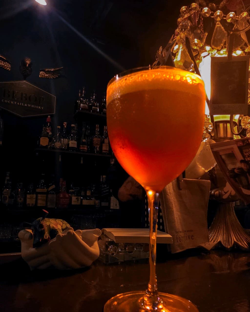 전남광주통합특별시 광산구 혼술 바 비밥 전남의 다찌석 위 전등 조명을 받는 주황빛 칵테일. 배경에 진열된 다양한 위스키 병들과 1인용 카운터 자리가 배치된 모습.