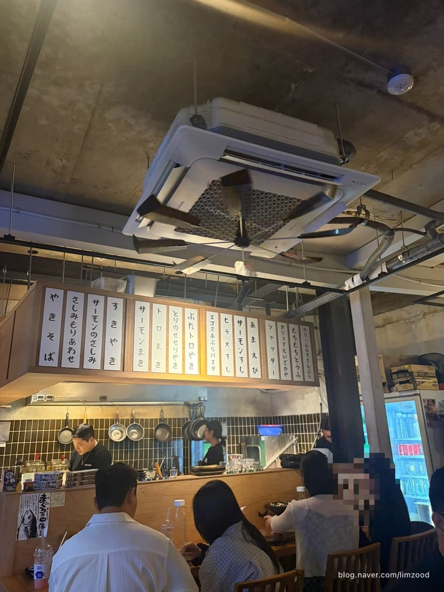 경상북도 경주시 혼술 이자카야 샷타 경북. 주황색 조명 아래 나무 다찌석과 일본어 메뉴판이 보이는 주방 구조. 1인석에 앉은 손님들과 요리하는 직원의 모습.