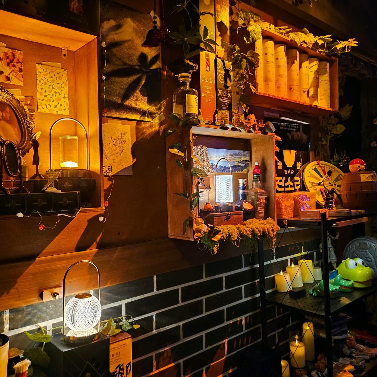 대전광역시 유성구 혼술바잔소리 봉명점 대전 바(Bar). 노란 조명이 비치는 목재 선반과 위스키 병, 1인 다찌석 주변에 배치된 장식품들이 어두운 벽돌 배경과 대비되는 혼술 장소.